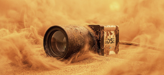 Caméra industrielle à moitié enfouie dans le sable, entourée de nuages de sable soulevés.