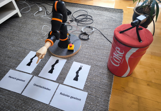 Le bras du robot pointe vers des feuilles de papier placées devant lui, la caméra IDS NXT se trouve à côté