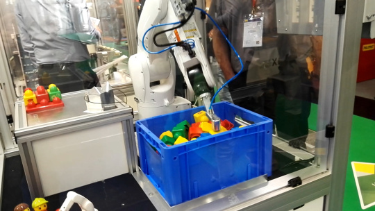 Un robot dans une cellule saisit une boîte bleue contenant des objets colorés