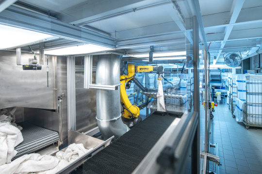 Un tapis roulant transportant du linge et un robot déployant le linge dans une blanchisserie industrielle.